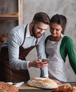Two bakers, a man and a woman, looking at a smartphone while working with freshly baked bread