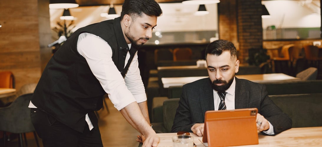 Restaurant manager reviewing business reports on a tablet, guided by a server, in a modern dining setting.