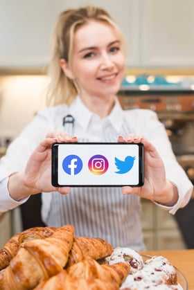 Smiling baker holding a smartphone displaying social media icons (Facebook, Instagram, Twitter) in front of assorted pastries